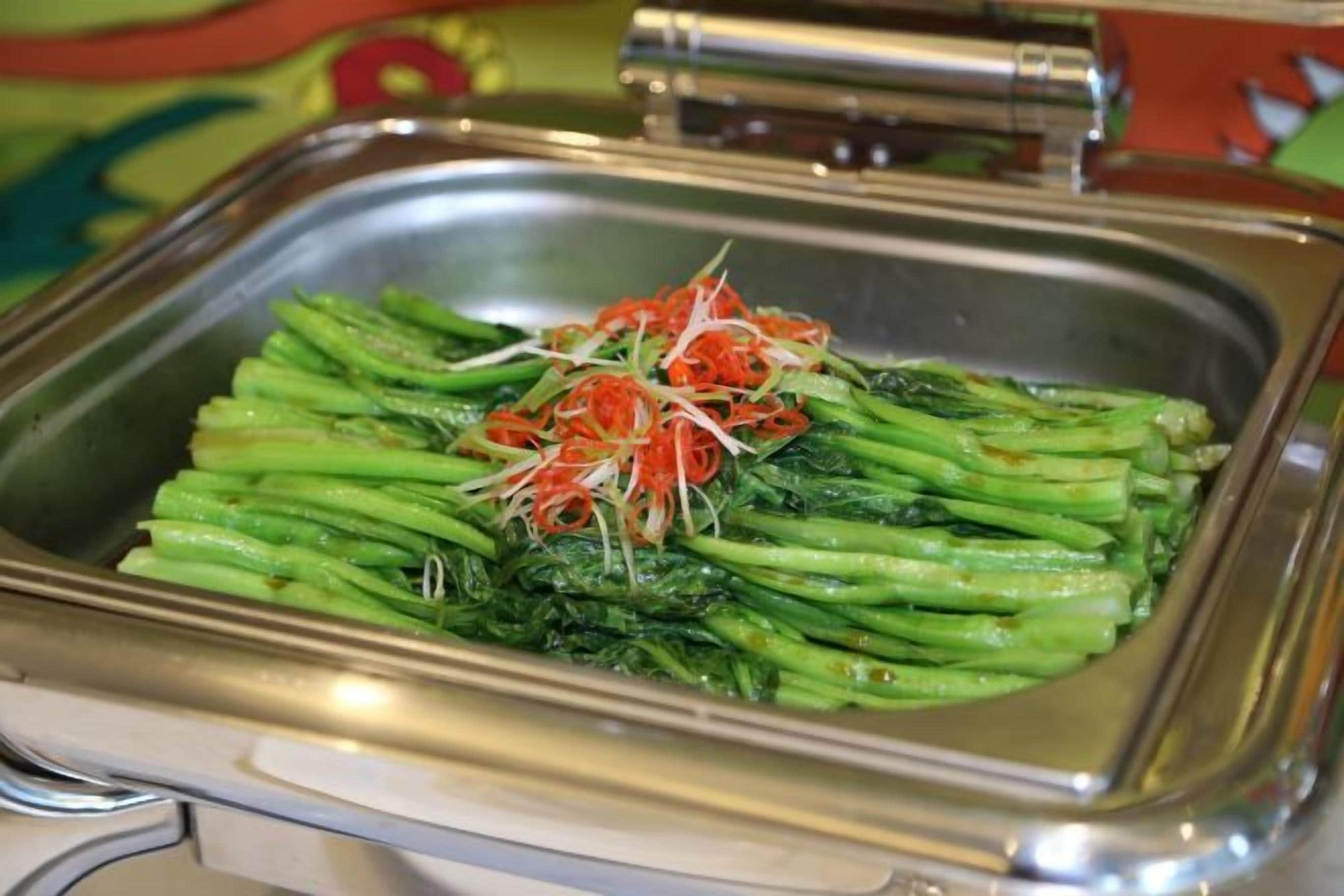 A buffet with a stainless steel serving dish containing green vegetables and shredded carrots.