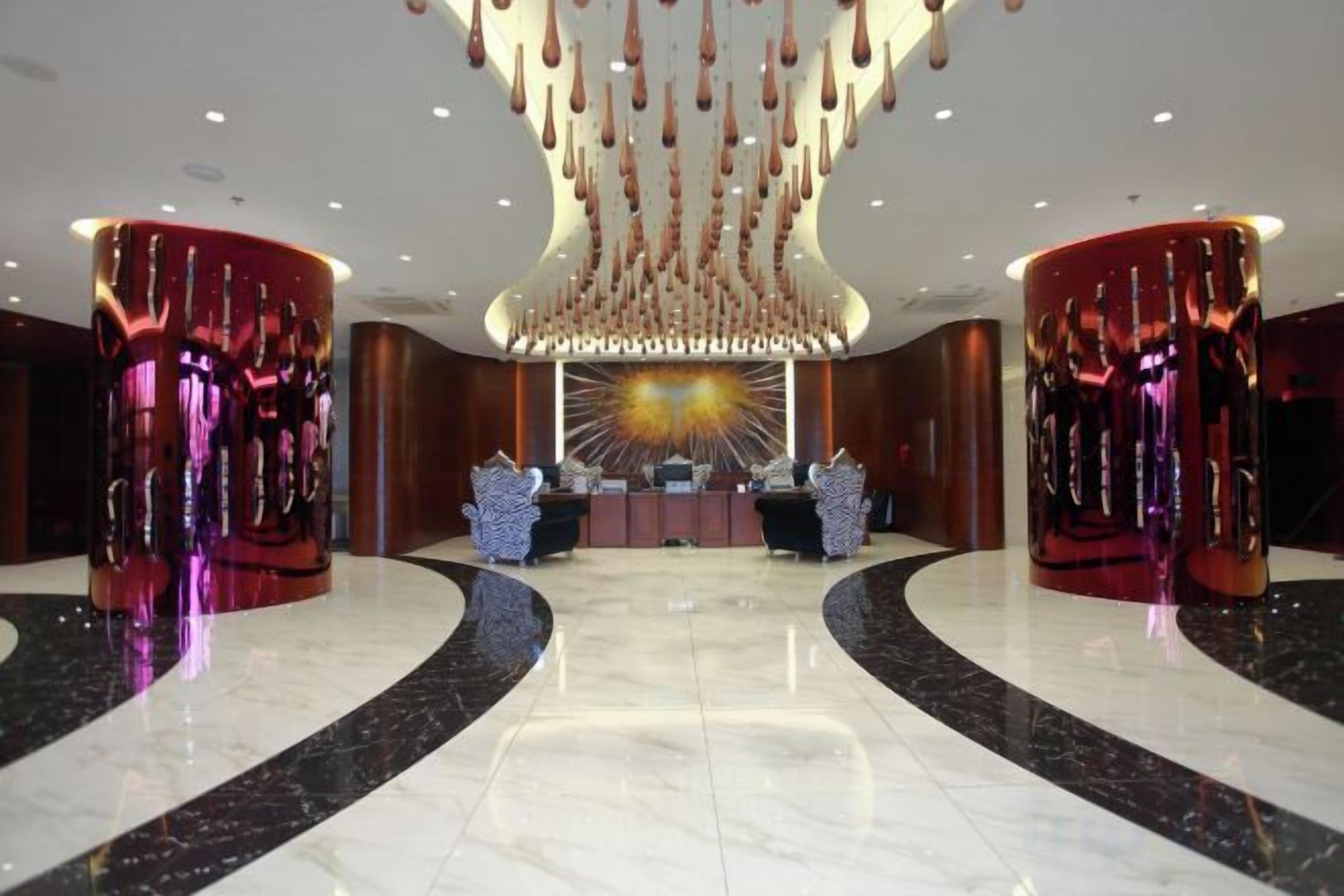 A spacious lobby with a central reception desk, two symmetrical red pillars, and a modern chandelier.