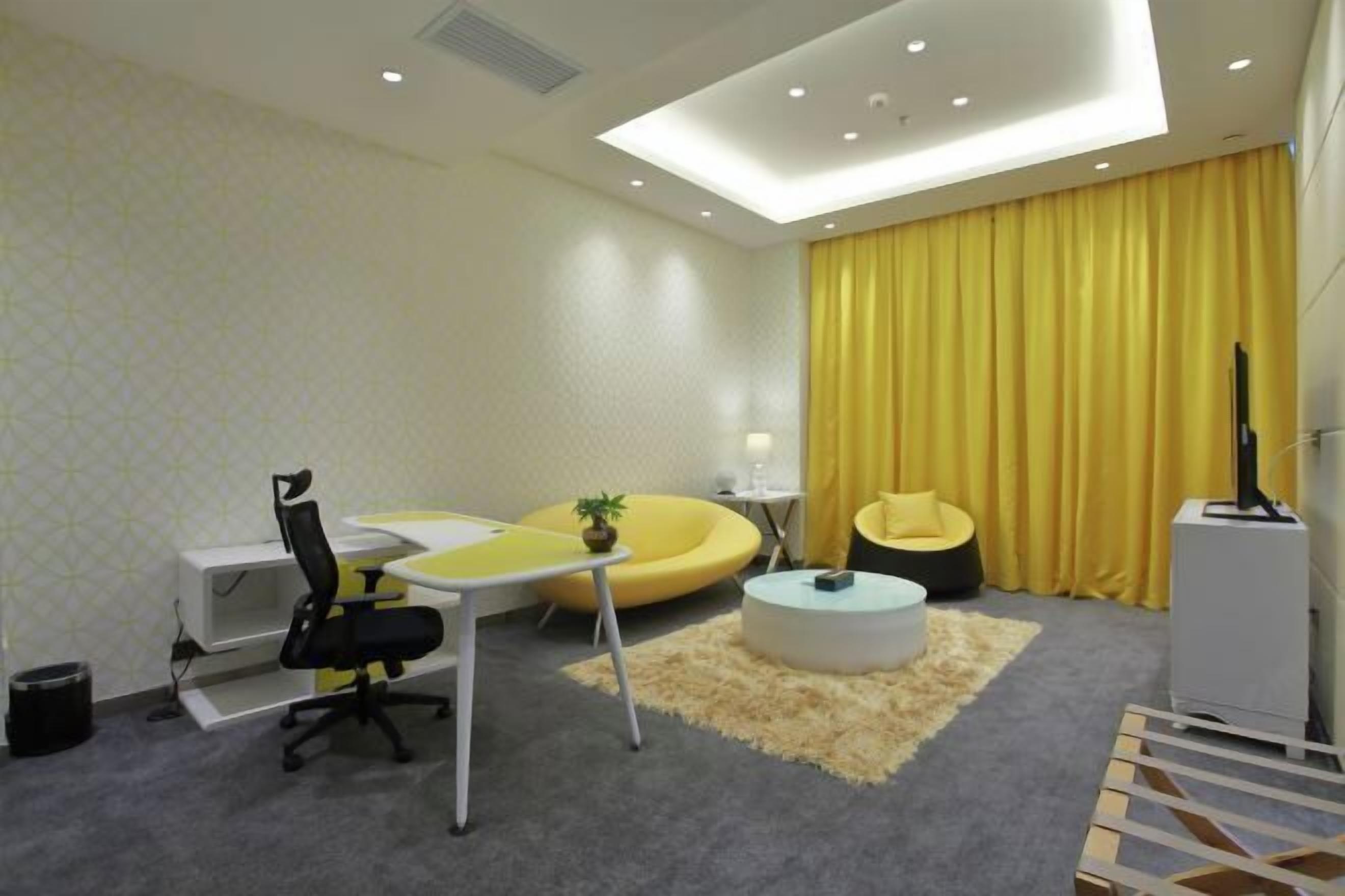 Un salon moderne avec un canapé jaune, une table basse ronde, un fauteuil de bureau noir et une petite plante.