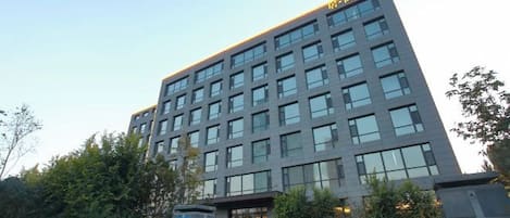 Un edificio de varios pisos con un diseño arquitectónico moderno, que cuenta con una entrada destacada y un estacionamiento con vehículos.
