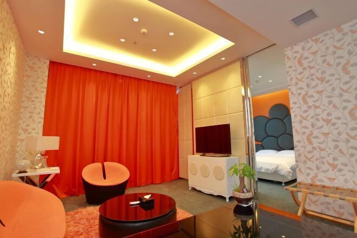 A modern hotel room with a large bed, a TV, a seating area with a round table, and a large window.