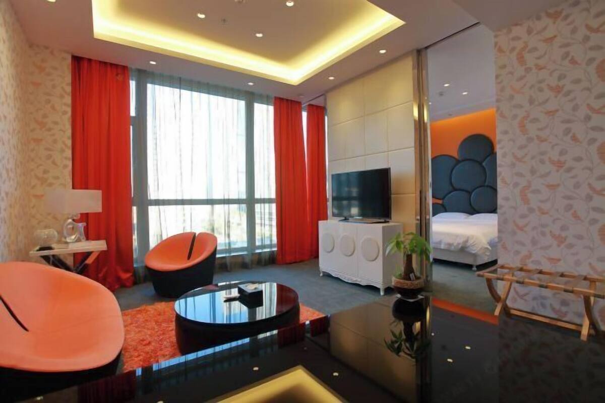 A modern hotel room with a flat-screen TV, a bed, a sofa, a coffee table, and a large window with curtains.