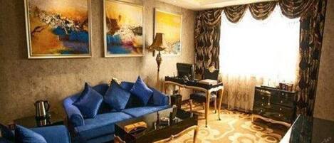 A modern living room with a blue sofa, a coffee table, and a desk. There are two paintings on the wall, a lamp, and a large window with curtains.