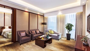 Una habitación de hotel con cama, dos sillones, un sofá, una mesa de centro con fruta y un ventanal con cortinas.
