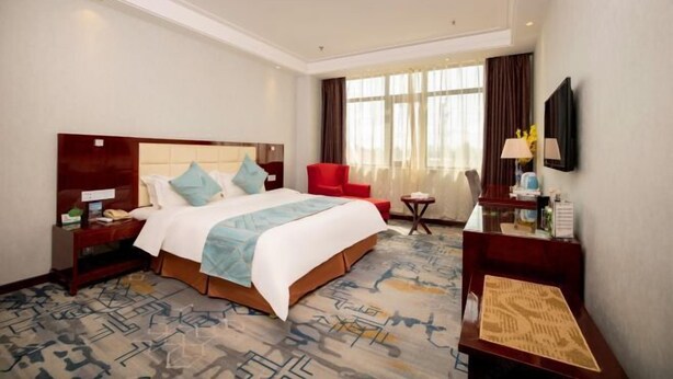 A hotel room with a large bed, a desk, a chair, a TV, and a window with curtains. - Zhongzhou Yihe Hotel (Xinxiang County)