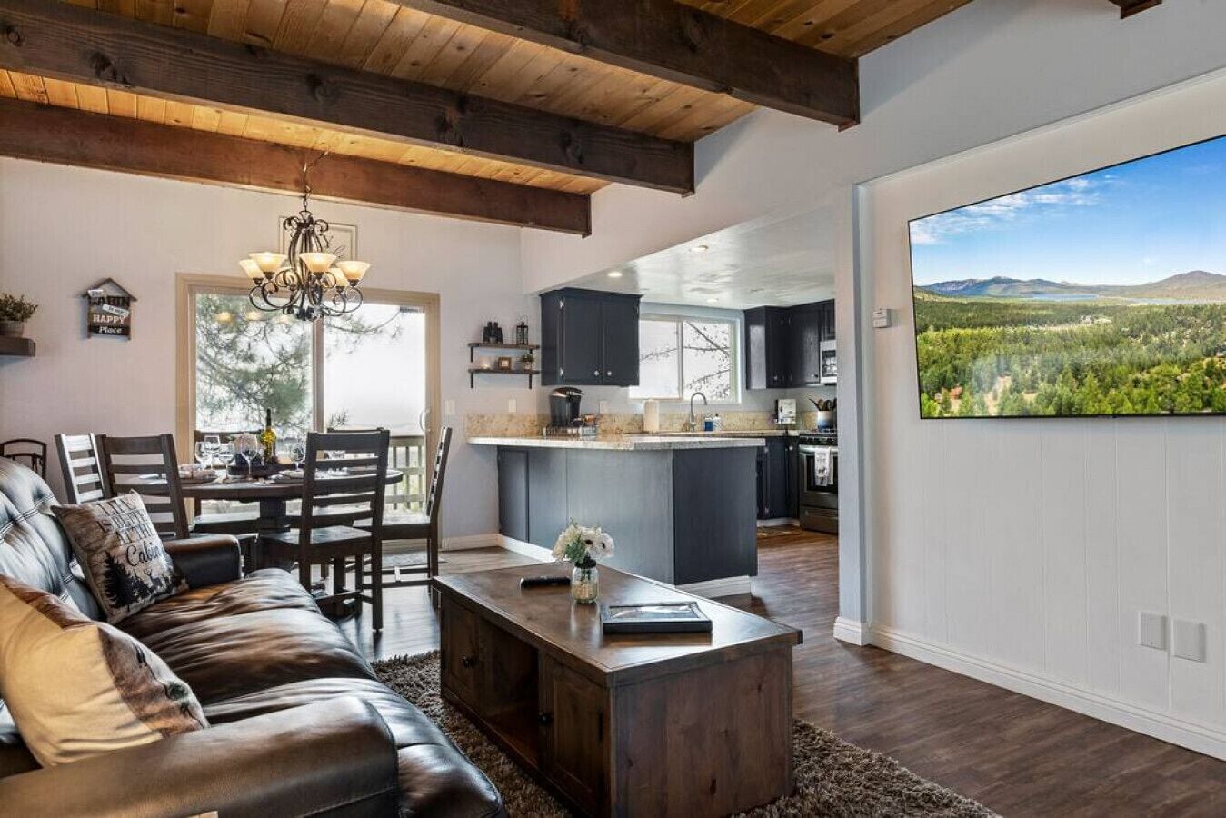 A spacious living room with a leather sofa, a wooden coffee table, and a dining area with chairs. A kitchen with dark cabinets and a large island is visible through an open doorway.