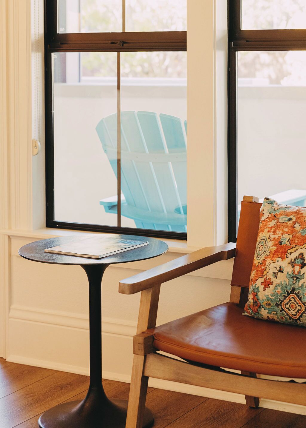 A room with a wooden chair and a small round table with a book. A window with a view of a blue chair outside is visible.