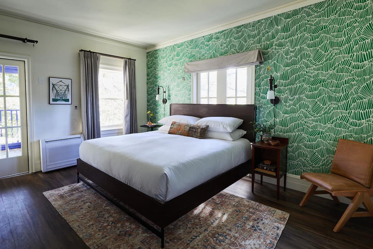 Un dormitorio con una cama grande, una silla, una mesita de noche, una ventana con cortinas, un radiador y una pared con estampado verde.