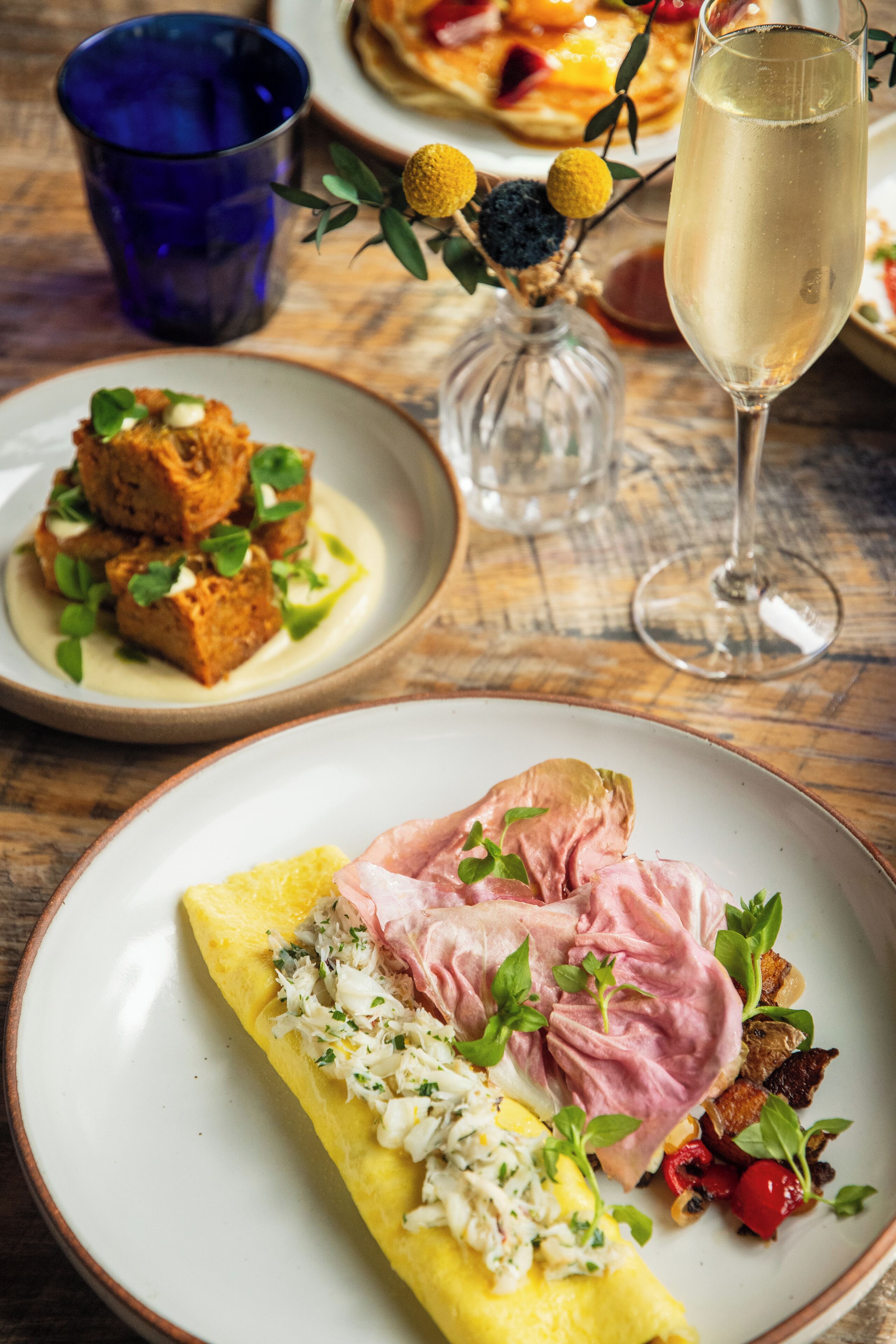 A dining table with various dishes, including a plate with a rolled omelette, a glass of champagne, and a blue glass.