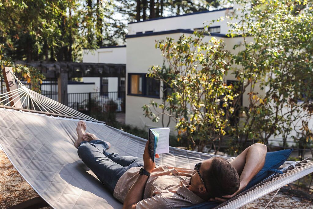 A hammock with two people, one reading a book, in a garden setting with a building in the background.