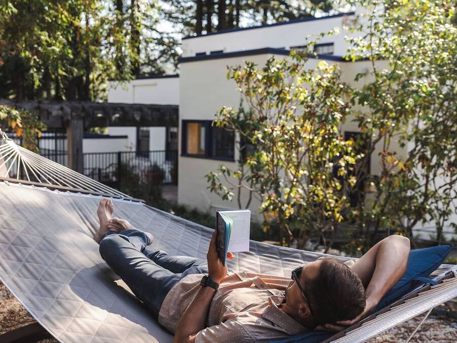 A hammock with two people, one reading a book, in a garden setting with a building in the background.