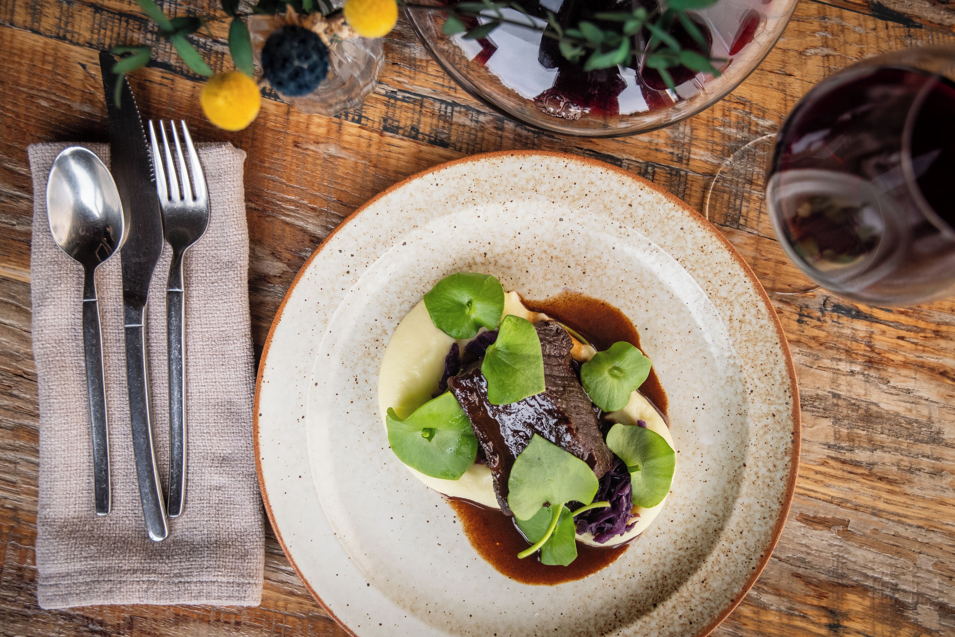 A plate of food with a fork and knife on a napkin, a glass of red wine, and a bowl of fruit on a wooden table.