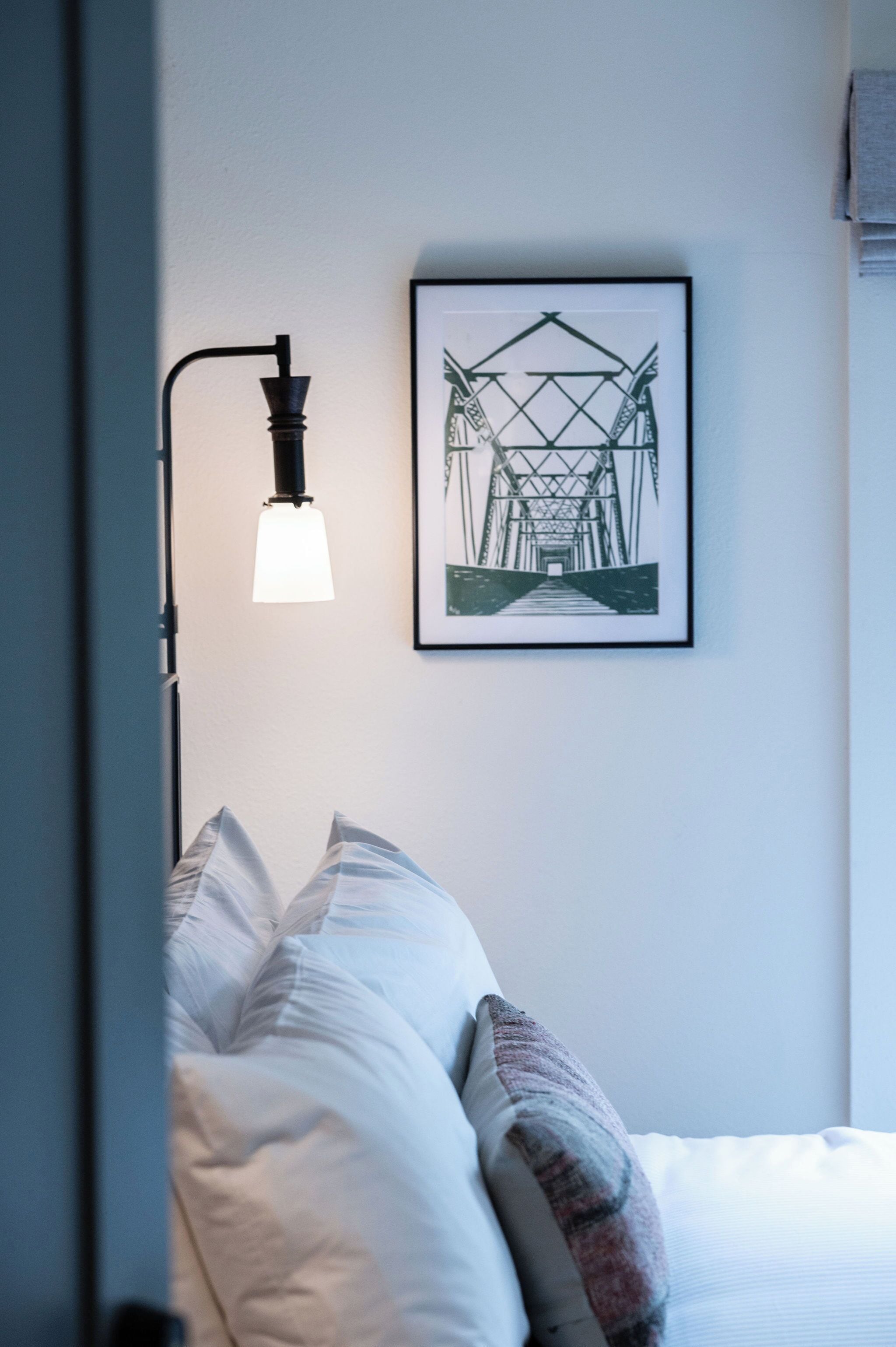 Quarto com cama, travesseiros, uma moldura na parede e uma luminária fixada na parede.