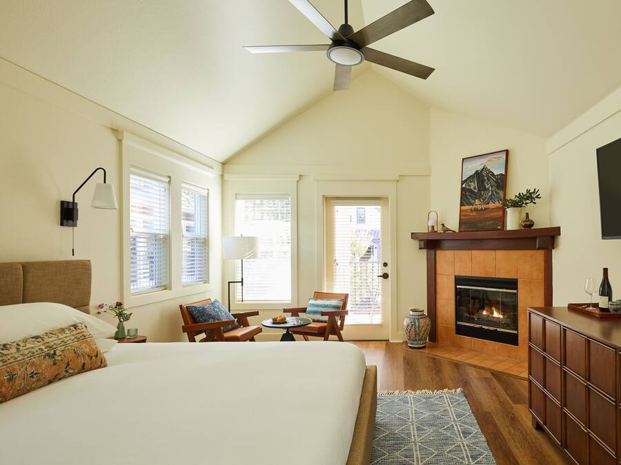 A bedroom with a large bed, a fireplace, a ceiling fan, and a dining area with chairs and a table.