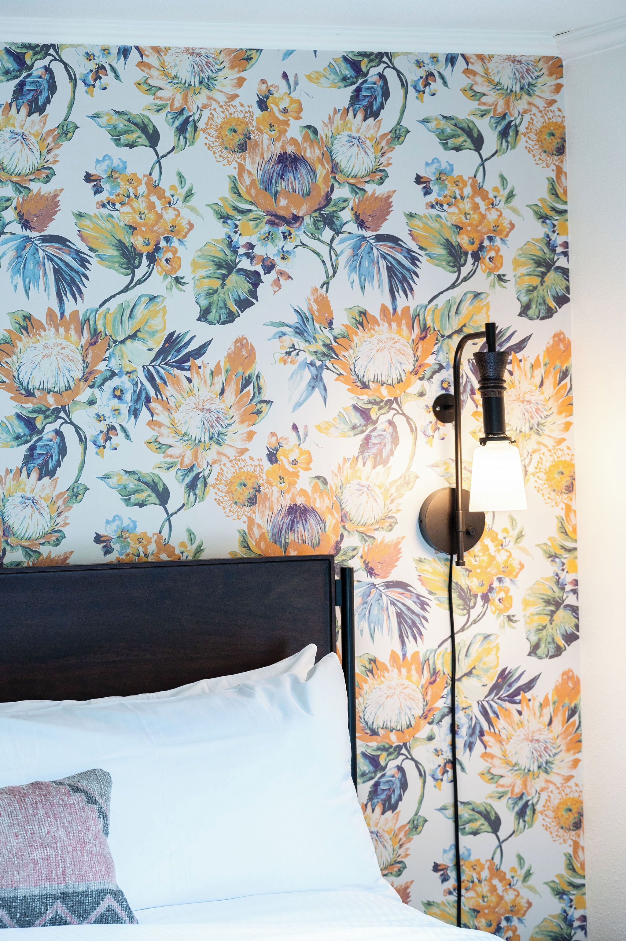Ein Bett mit weißen Laken und einem einzelnen Zierkissen, platziert an einer Wand mit blumigem Tapetenmuster.