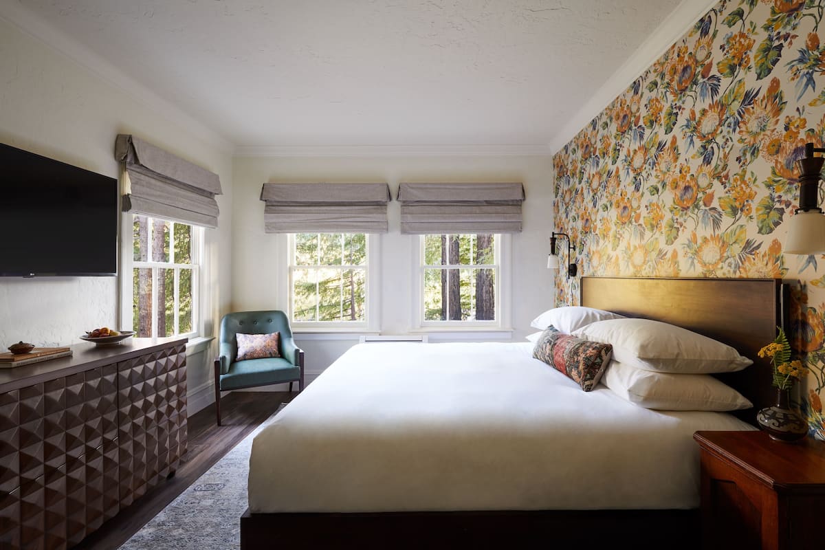 Un dormitorio con una cama grande, un televisor montado en la pared y una ventana con vistas a árboles.