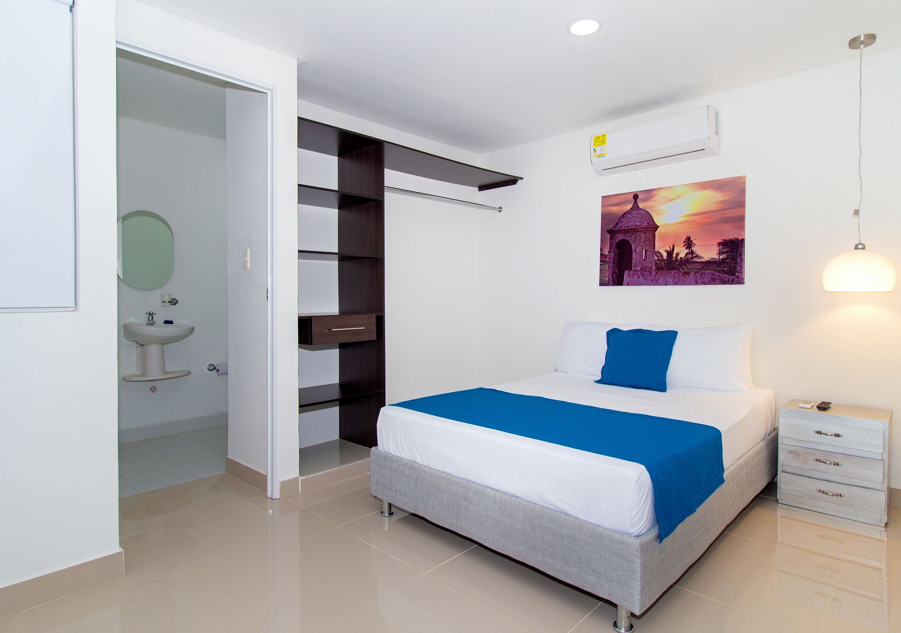 A modern bedroom with a bed, a nightstand, a wall-mounted air conditioner, a painting, and a bathroom with a sink and mirror.