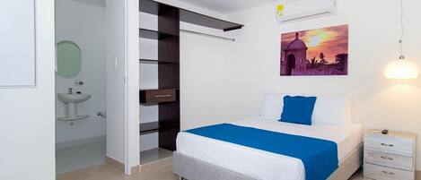 A modern bedroom with a bed, a nightstand, a wall-mounted air conditioner, a painting, and a bathroom with a sink and mirror.