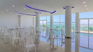 A spacious indoor area with rows of white plastic chairs arranged for seating, large windows offering a view of the sea, and a modern ceiling design with recessed lighting. - Hotel Playa Norte (Cartagena)