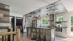 A modern kitchen with a wooden accent wall, a central island with stools, stainless steel appliances, and pendant lighting. - Le Panaché - Cottage to rent in Stoneham (CITQ: 298070) (Stoneham-et-Tewkesbury)