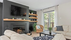 A modern living room with a fireplace, a television mounted on the wall, and a balcony with a view of trees. - Le Panaché - Cottage to rent in Stoneham (CITQ: 298070) (Stoneham-et-Tewkesbury)