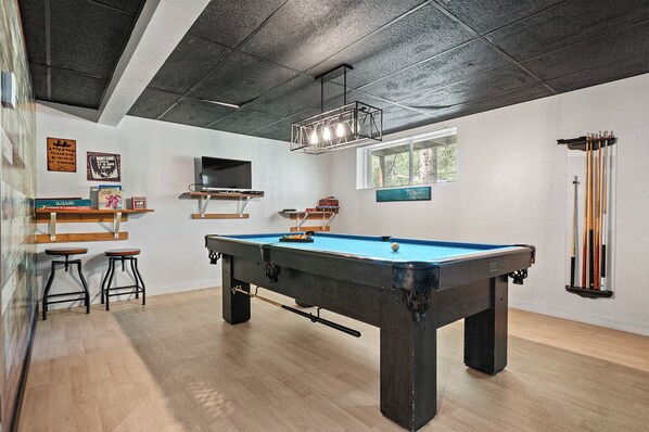 A pool table in a room with wooden flooring, a ceiling with exposed beams, and a window with blinds. - Le Panaché - Cottage to rent in Stoneham (CITQ: 298070) (Stoneham-et-Tewkesbury)