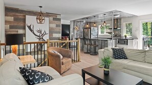 A modern living room with a sofa, armchair, and coffee table. A kitchen with a bar area and a dining table is visible in the background. - Le Panaché - Cottage to rent in Stoneham (CITQ: 298070) (Stoneham-et-Tewkesbury)