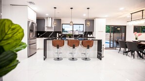 A modern kitchen with a central island, three bar stools, stainless steel appliances, pendant lighting, and a view to the outdoors.