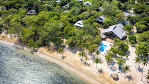An aerial view of a coastal resort with multiple bungalow-style accommodations surrounded by dense tropical vegetation, a sandy beach, and a clear blue pool area. - Laru Beya Palmetto Bay (Roatan)