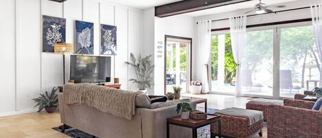 A spacious living area with a sofa, armchairs, and a TV. There are potted plants and a ceiling fan.