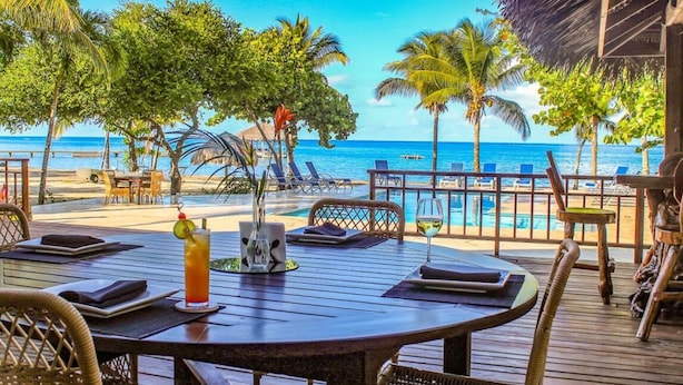 A wooden deck with tables and chairs, overlooking a pool and beach with palm trees and a clear blue sky. - Laru Beya Palmetto Bay (Roatan)