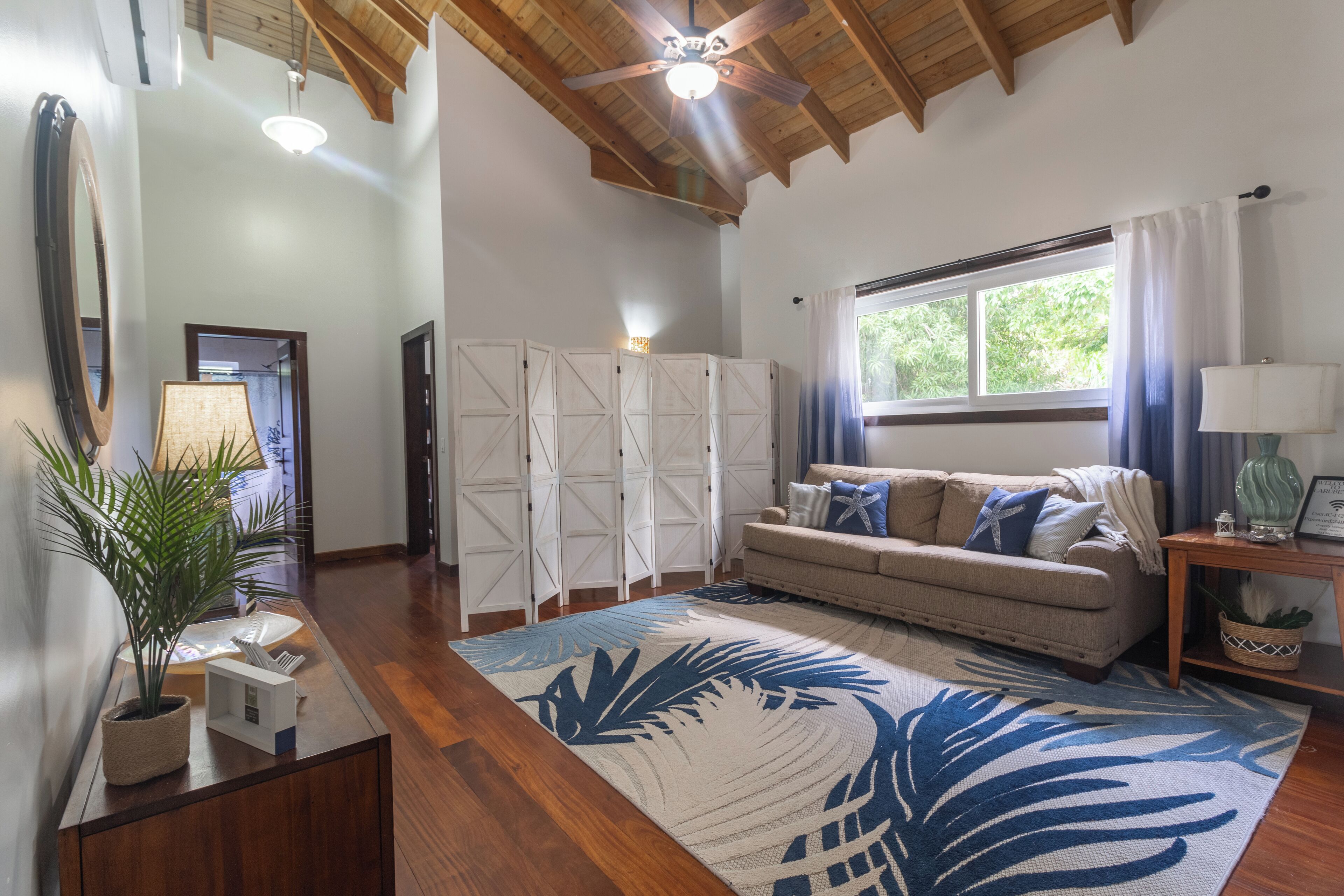A spacious living room with a large window, a ceiling fan, and a wooden floor.