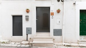 Un edificio con un acceso amplio, una ventana pequeña y una puerta verde. Hay dos plantas en macetas en la pared.