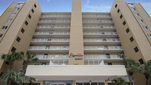 Ein mehrstöckiges Hotel mit einem zentralen Eingang, Balkonen in jeder Etage und Palmen im Vordergrund.