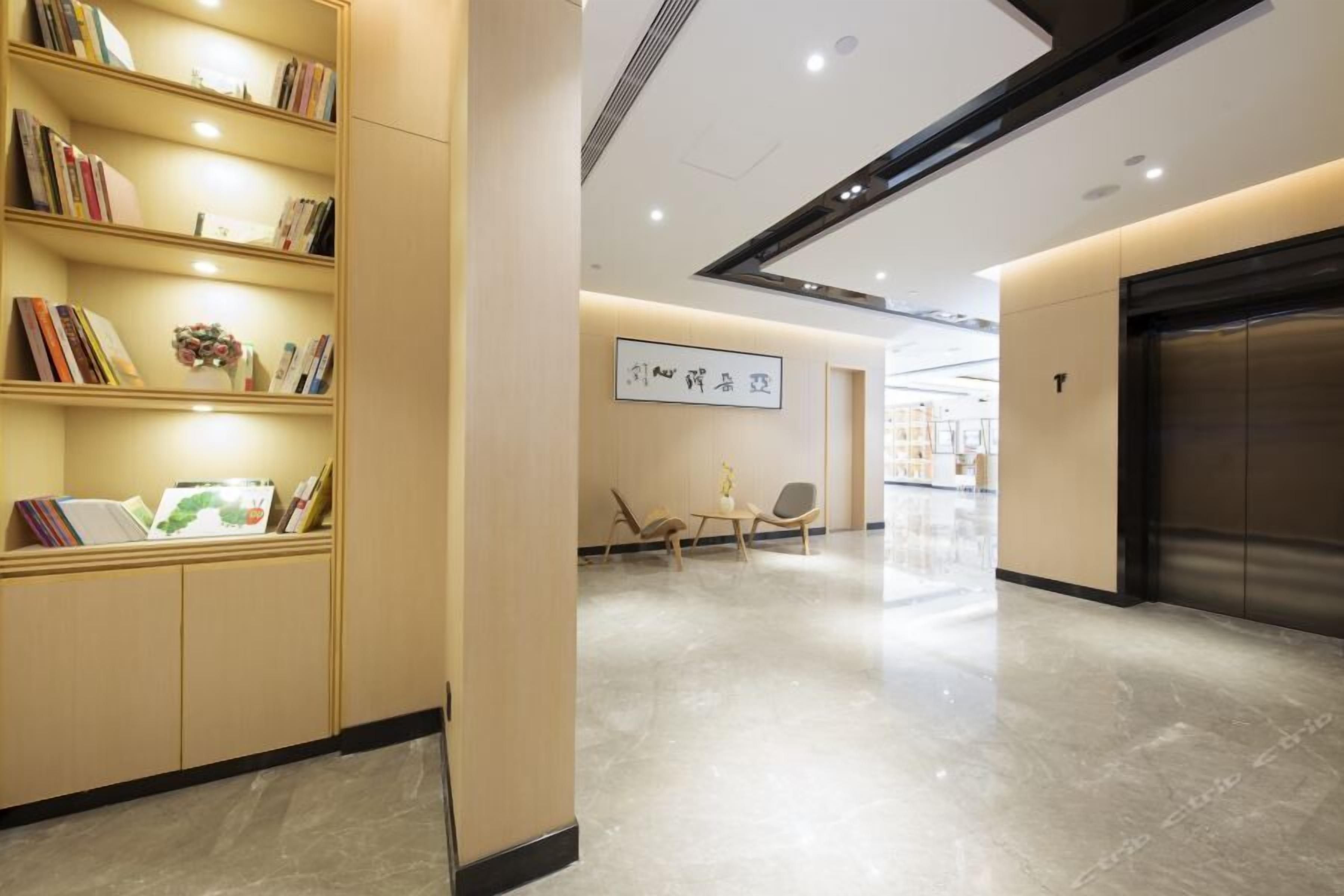 Um hall de hotel moderno com piso de mármore, uma estante com livros e um vaso, uma área de estar com duas poltronas e um elevador.