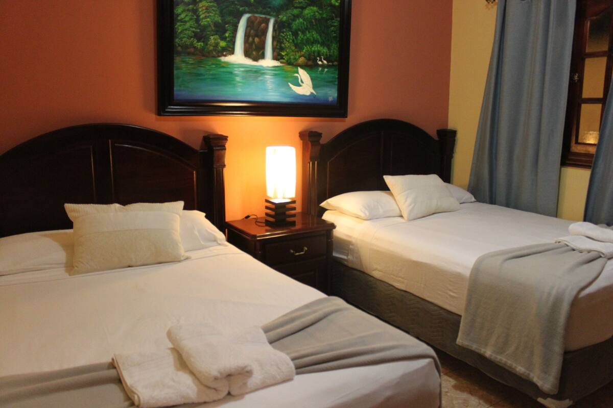 Zwei nebeneinanderliegende Betten mit weißen Bettwäsche, ein Nachttisch mit Lampe und ein Bild eines Wasserfalls an der Wand.