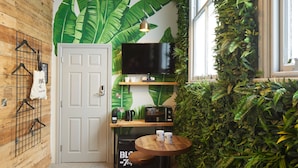 Una habitación compacta con un panel de madera, un mural grande con una hoja verde, un televisor, una máquina de café y una pequeña mesa redonda con taburetes.