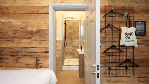 Habitación de hotel con pared de madera, cama, zona de ducha, perchero y bolsa para secador.