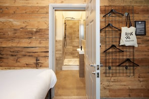 A hotel room with a wooden wall, a bed, a shower area, and a rack with hangers and a hairdryer bag. - Blok-74 (Brighton)