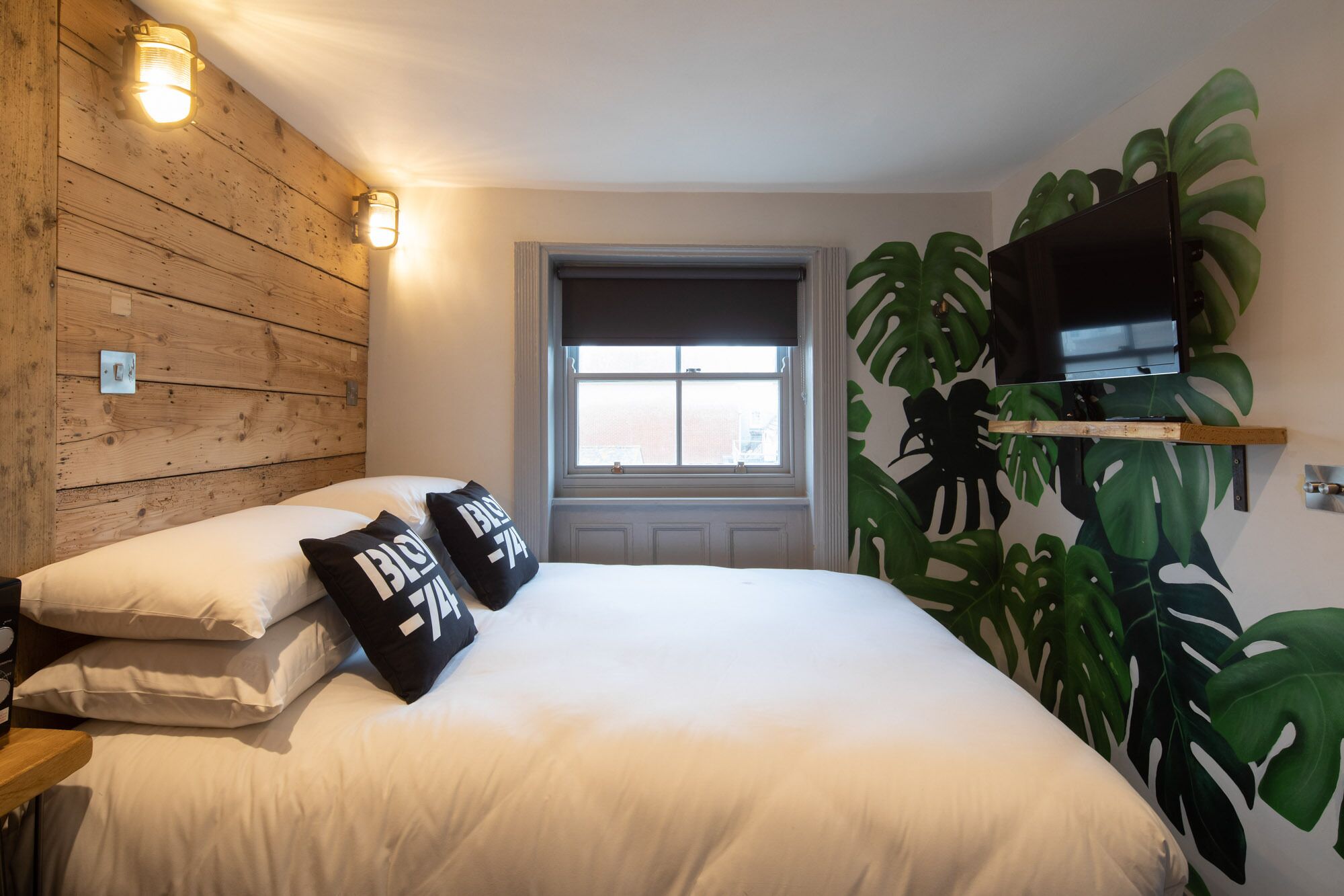 A bedroom with a wooden wall, a window, a bed with white bedding, two black pillows with text, a mounted TV, and a large plant.