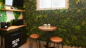 Un café acogedor con una pared cubierta de follaje verde, una barra de madera con máquinas de café, dos taburetes y una mesa pequeña con tazas.