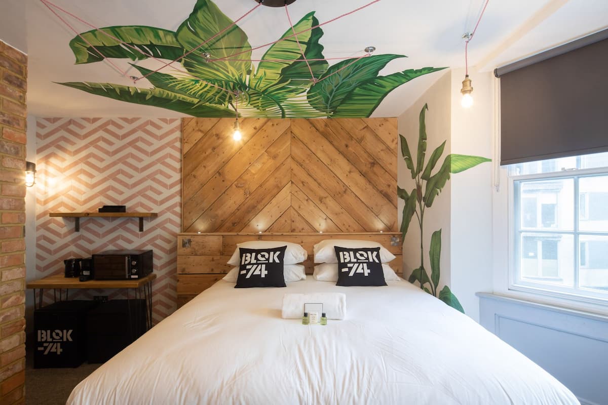 Ett hotellrum med en sänggavel i trä med fiskbensmönster, en säng med vita sängkläder, två kuddar med texten ”BLOK 74”, ett litet bord med en kaffemaskin och en stor växt som hänger från taket.