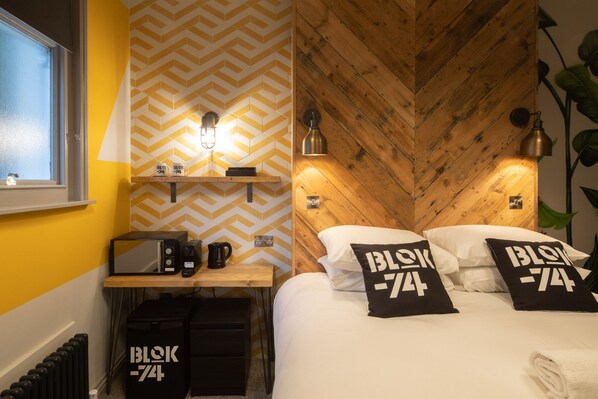 A hotel room with a chevron-patterned wall, wooden headboard, and two bedside lamps. There are two black pillows with 'BLOK-74' text, a coffee maker, and a radio on a wooden shelf. - Blok-74 (Brighton)