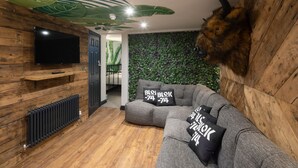 Una habitación acogedora con piso de madera, un sofá modular gris con cojines negros, un televisor montado y una pared con un estampado de hojas verdes.