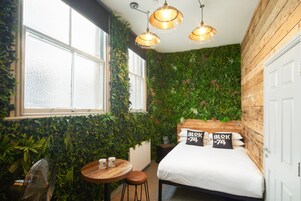 Una habitación con una cama, una mesita y una silla. Hay tres lámparas colgantes sobre la cama. Las paredes están cubiertas de plantas verdes.