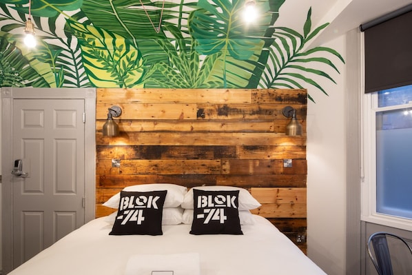 Un dormitorio con cabecera de madera, dos lámparas fijadas a la pared y una cama con ropa de cama blanca y dos almohadas con el texto 'BLOK 74'.