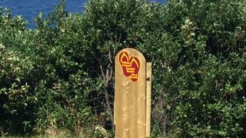 A wooden gate with a circular sign featuring a logo and text, surrounded by dense green foliage.