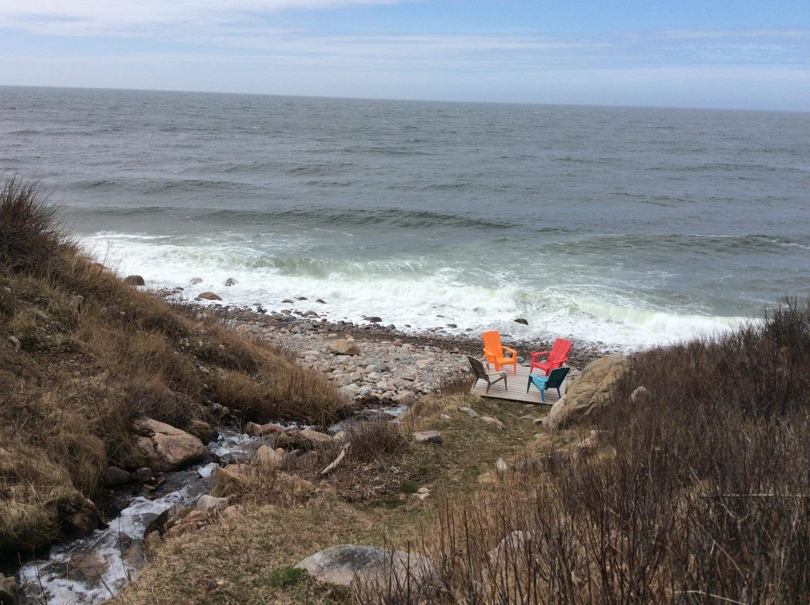 Een rotsachtige kustlijn met golven die op het strand breken. Drie stoelen staan op een houten platform met uitzicht op de oceaan.