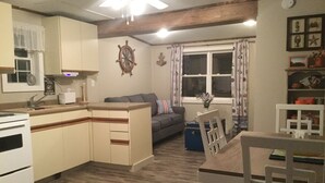 A compact kitchen with a central island, a dining area with a wooden table and chairs, a gray sofa, and a window with curtains.