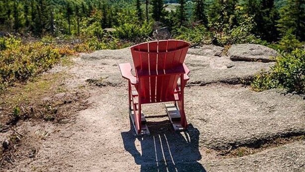 A red Adirondack chair on a rocky outcrop overlooking a forested valley with a view of the ocean and distant landforms.